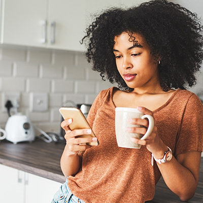 Ung kvinna står och dricker kaffe och tittar på sin telefon i ett kök.
