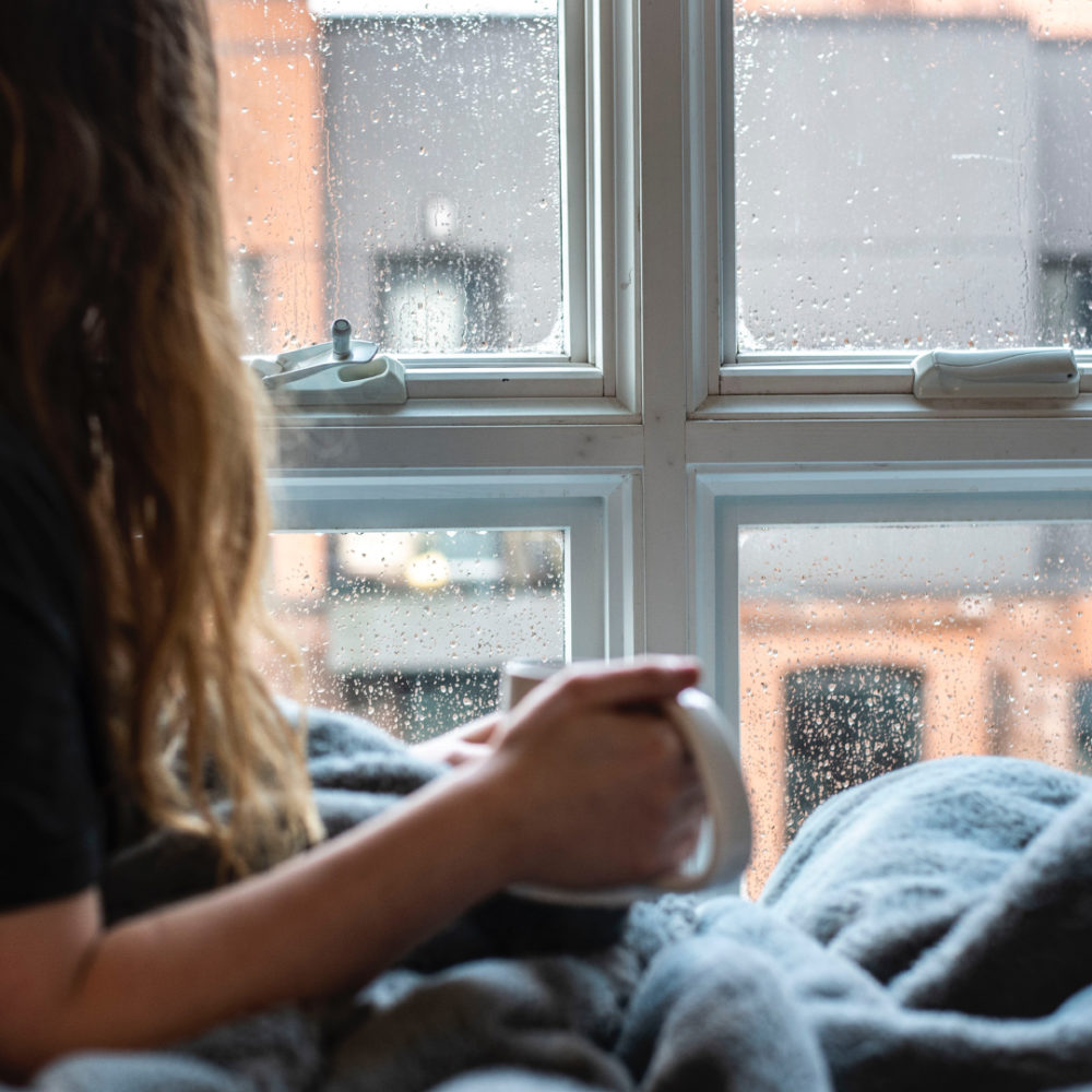 Person med långt hår och kaffemugg i handen sitter med filt över knäna och tittar ut genom regningt fönster.